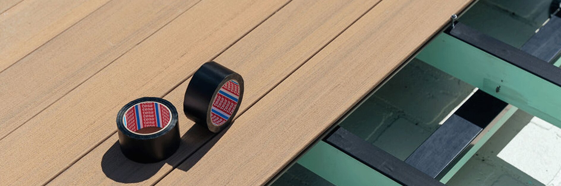 Two rolls of black tesa tape with red and blue patterns are placed on a wooden deck surface. The deck is partially constructed with visible beams underneath. Sunlight casts shadows on the deck. (This text has been generated by AI)