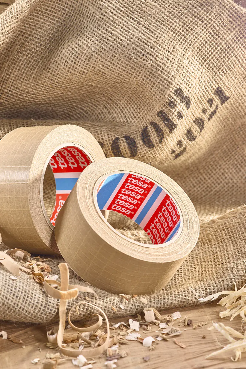 Two rolls of beige tesa tape with red inner labels sit on a burlap sack. Wood shavings are scattered around, with some curly pieces nearby. A wooden surface is in the background, and soft light illuminates the scene from the upper right. (This text has been generated by AI)