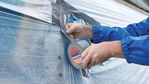 Hands apply a strip of tesa tape to a clear plastic sheet, securing it in place. The person is wearing a blue jacket.