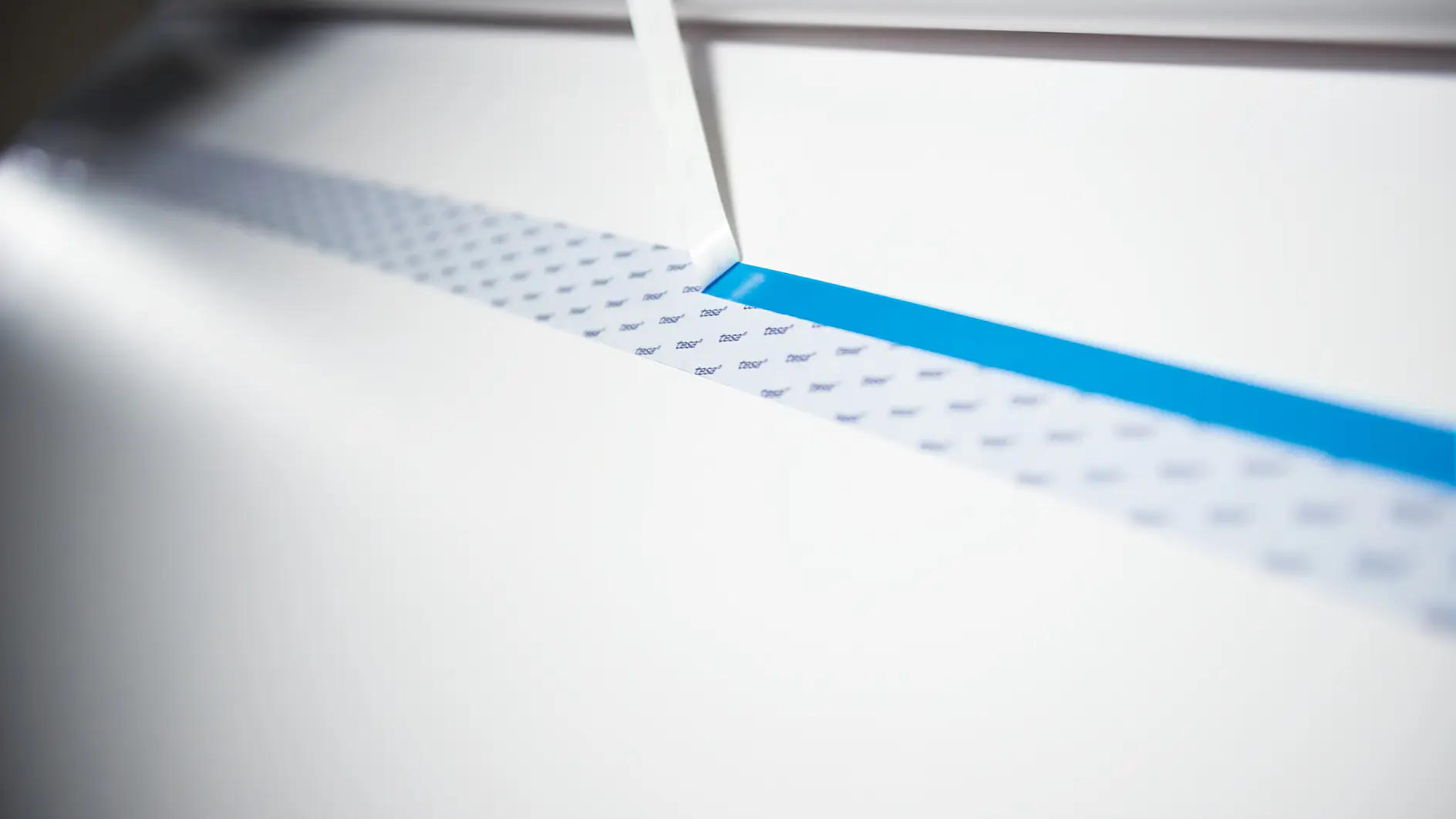 A close-up of a white material being cut by a thin blade. The material features a repeating pattern of small, dark tesa tape logos and a blue stripe running parallel to the cut.