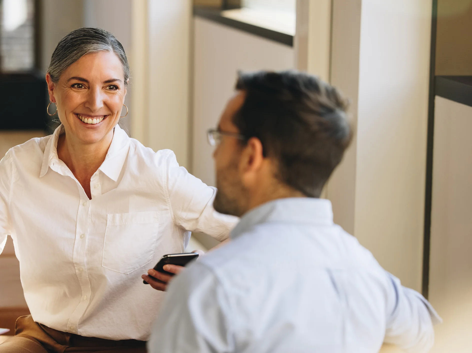 tesa-IGM_CollaborationPage_ContactForm-788x442 A woman with gray hair is smiling and holding a smartphone, sitting across from a man wearing glasses. Both are dressed in casual business attire and sitting in an office environment with large windows. (This text has been generated by AI)