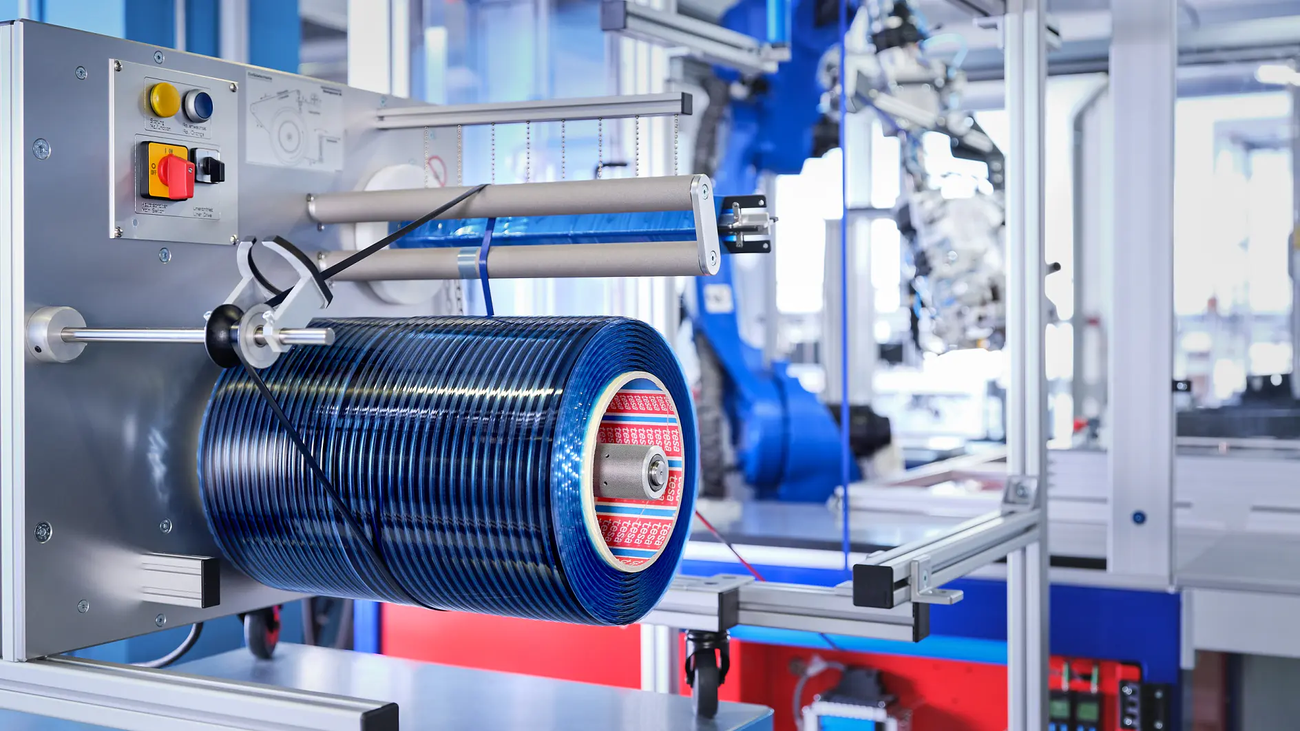 A manufacturing facility with industrial equipment, featuring a blue cylindrical component mounted on a metal frame using tesa tape. The machinery includes buttons, wires, and a robotic arm in the background. The setting is clean and organized.