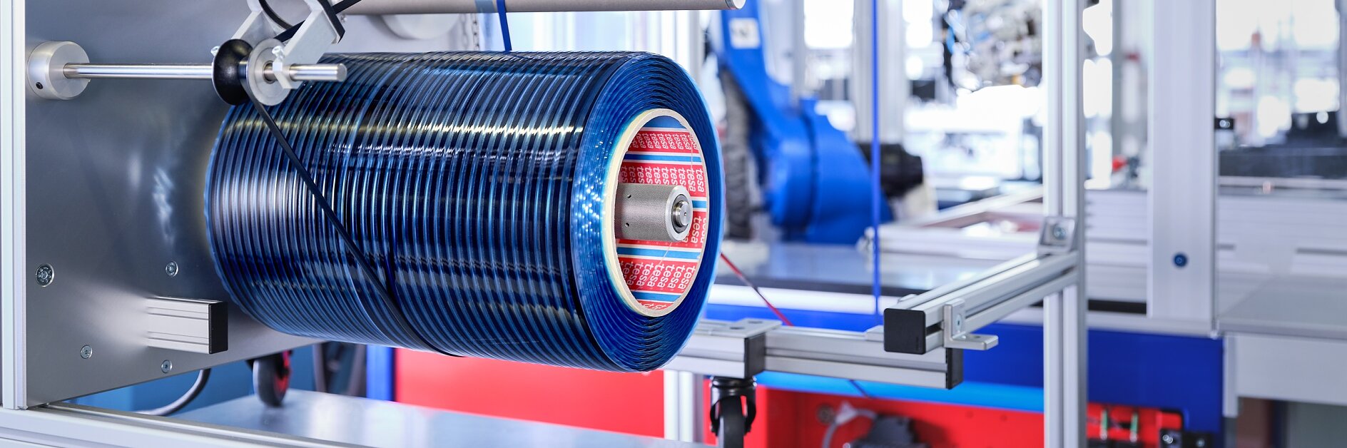 A manufacturing facility with industrial equipment, featuring a blue cylindrical component mounted on a metal frame using tesa tape. The machinery includes buttons, wires, and a robotic arm in the background. The setting is clean and organized.