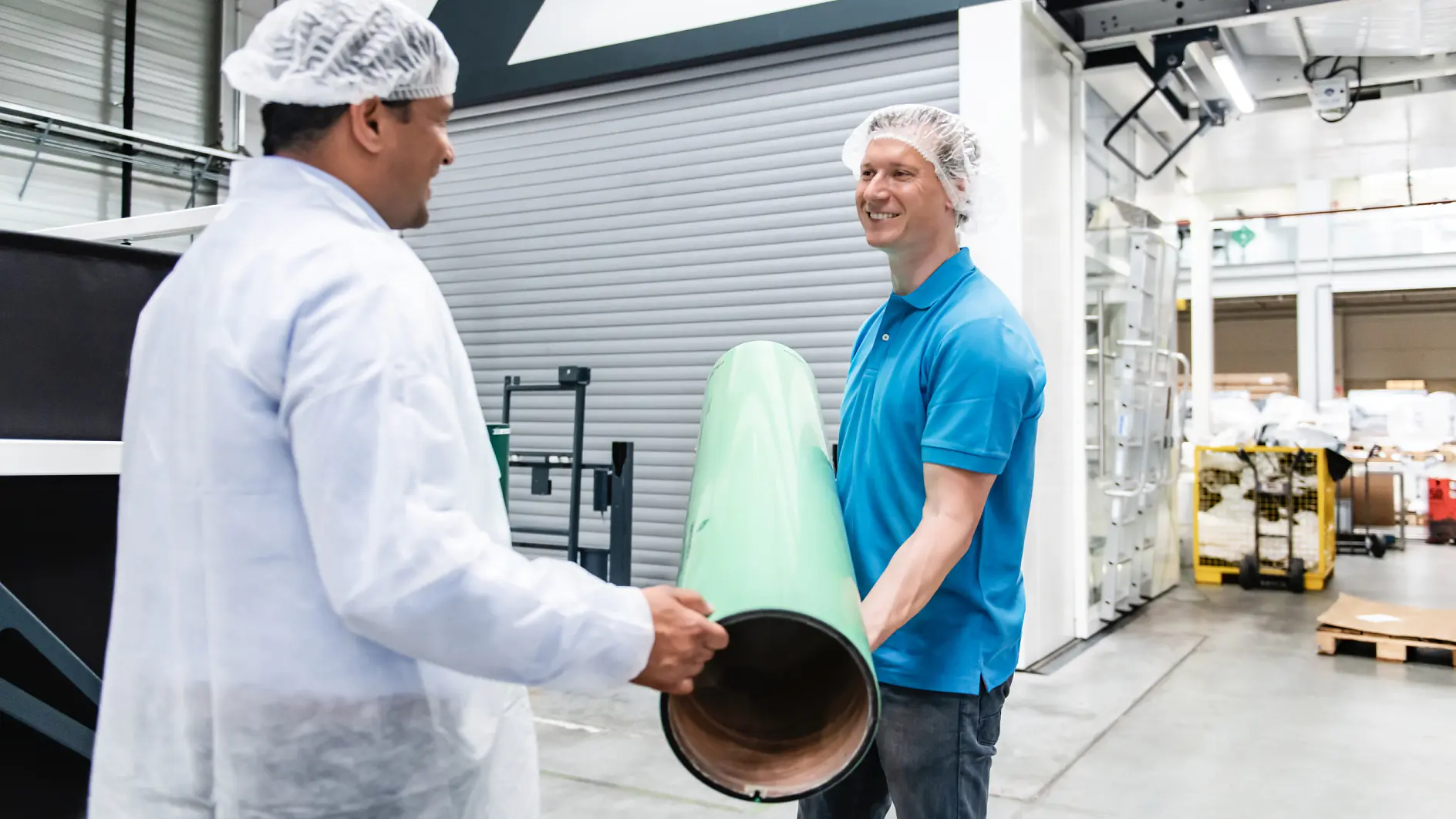 Two men wearing hairnets are in a manufacturing facility. One, dressed in a blue polo shirt, is holding a large green cylindrical object, while the other man, in a white lab coat, observes. Machinery and equipment can be seen in the background.