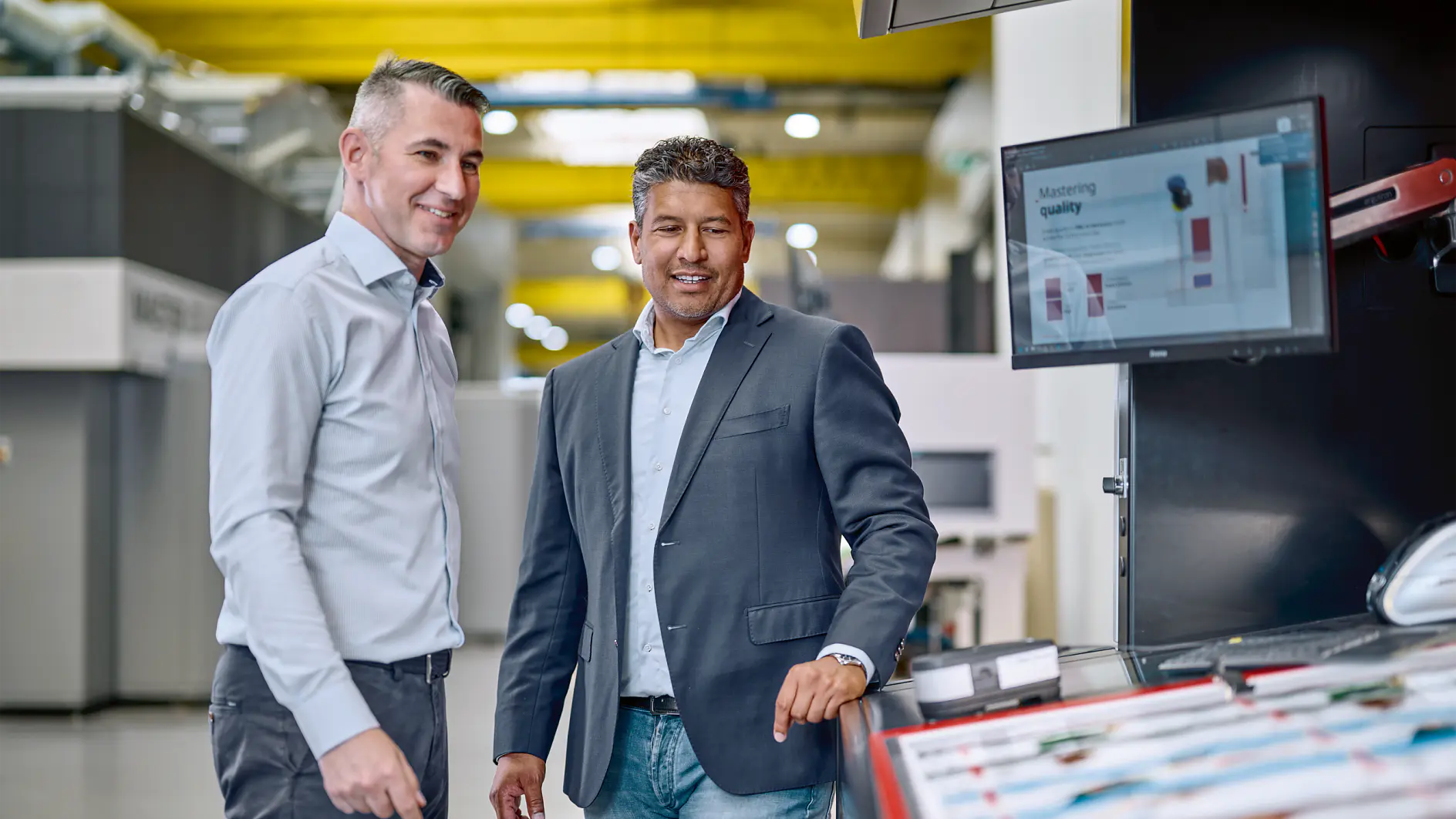 Two men in business attire stand in an industrial setting. One man is pointing at a computer screen displaying data, while the other smiles. They are surrounded by machinery and equipment, including several rolls of tesa tape. (This text has been generated by AI)