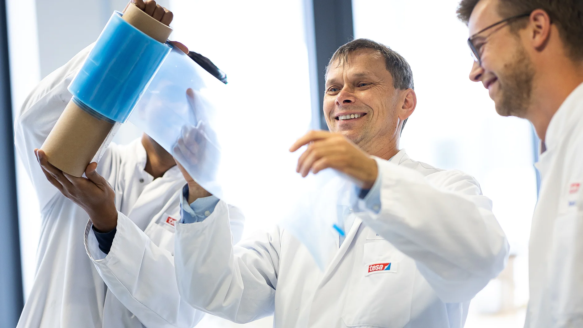 Three people in lab coats examine a roll of transparent blue tesa tape in a bright laboratory setting. One person holds the tesa tape upright, while another pulls a section away. They appear focused and engaged. (This text has been generated by AI)