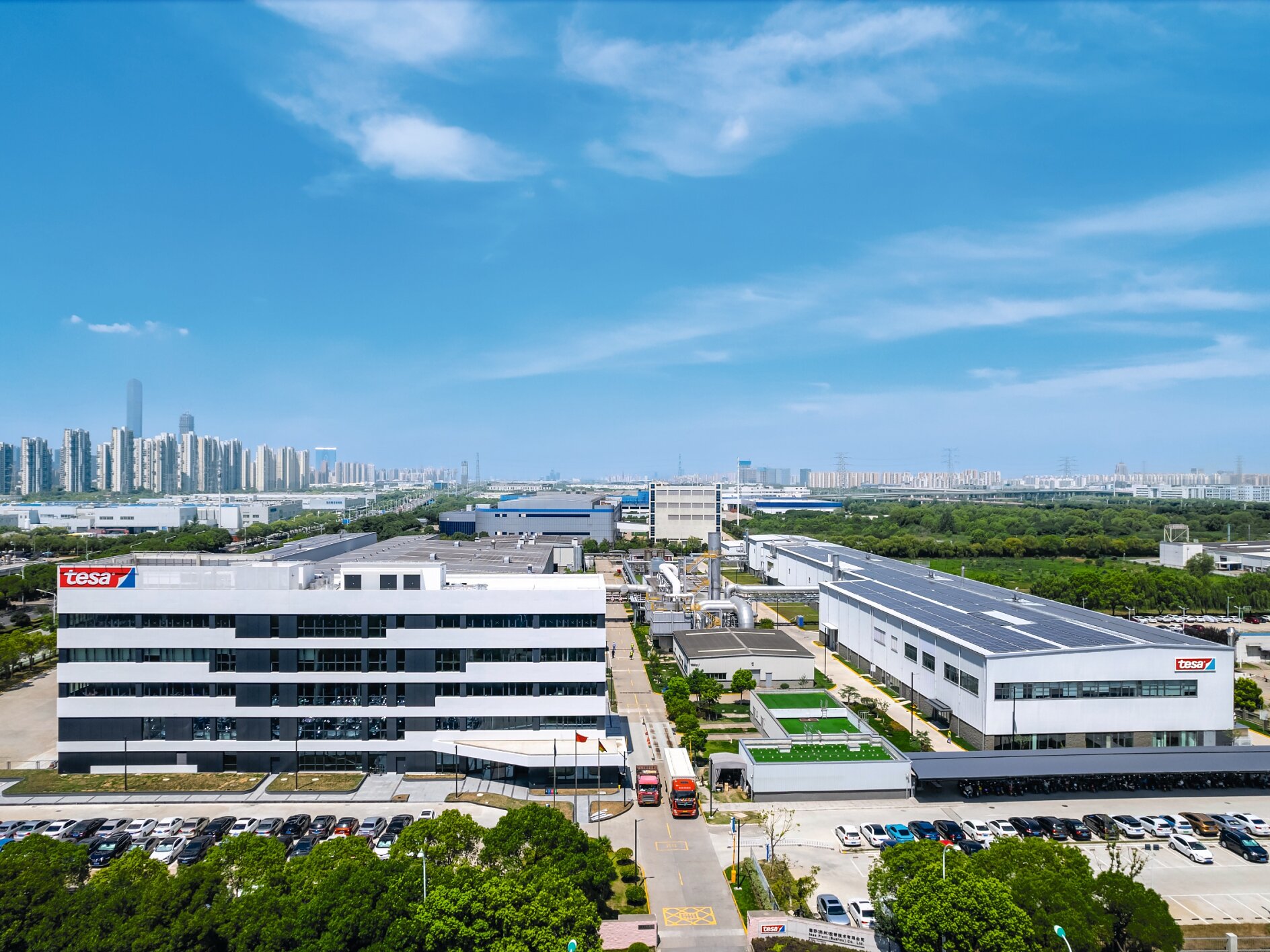 Aerial view of a sprawling industrial complex with multiple white buildings under a blue sky. The complex features parking areas and roads. A city skyline is visible in the distance. (This text has been generated by AI)