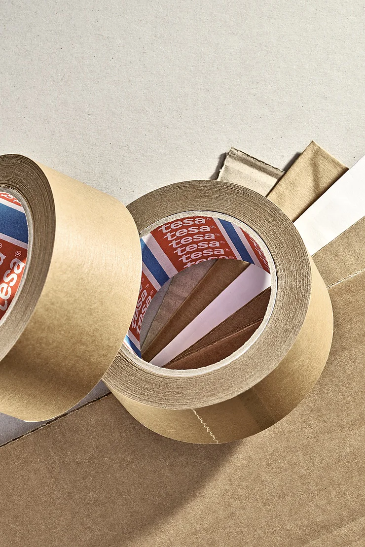 4513-superior-paper-packaging-tape-mp (3) Two rolls of brown tesa tape are shown on a light surface. One roll is upright with the red, white, and blue inside label visible. The other roll is partially unrolled, with its adhesive side facing up. Nearby are sheets of cardboard. (This text has been generated by AI)