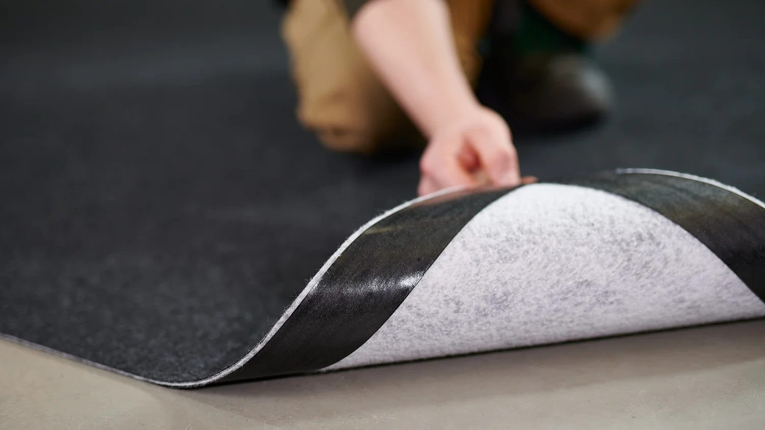 5938_2806_72dpi A person wearing brown pants is shown installing a black and white carpet tile over a smooth, gray floor. The focus is on the persons hand lifting the edge of the tile, revealing the underside secured with tesa tape. (This text has been generated by AI)