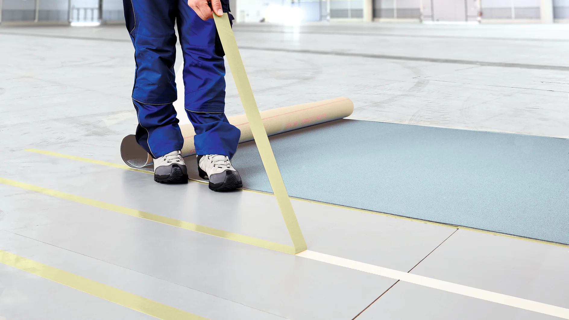 tesa_5939_step7of7_0126_300dpi-cms A person in blue pants and white sneakers is unrolling a large sheet of blue paper on a gray floor. They hold a roll of yellow tesa tape, applying it across the floor to create parallel lines. The background shows an empty, spacious room. (This text has been generated by AI)