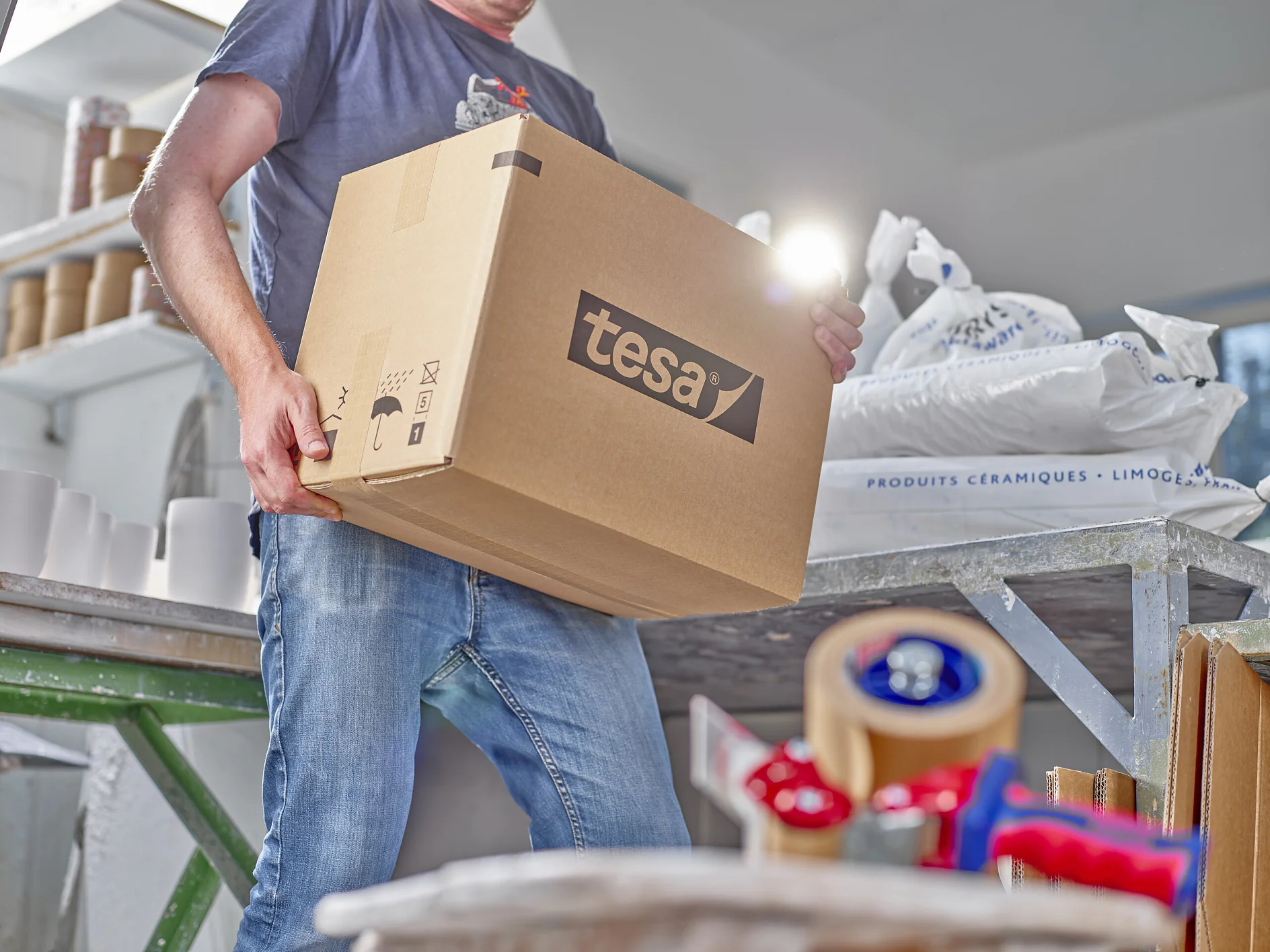 tesa-60013-self-adhesive-reinforced-paper-packaging-tape-17-ap-cms A person holds a large cardboard box labeled tesa in a workspace. Various packing materials are scattered around, including tesa tape dispensers and white sacks. The person is wearing casual clothing, with jeans and a dark top. (This text has been generated by AI)