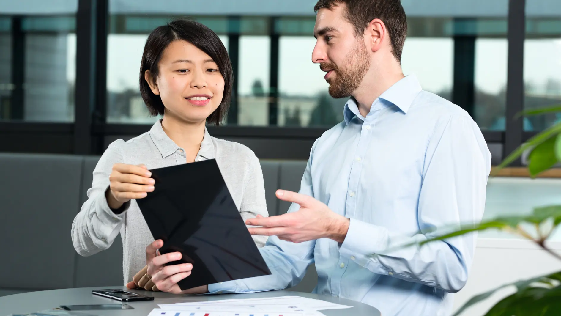 A man and a woman are sitting at a round table, engaged in discussion. The woman holds a tablet, showing it to the man, who gestures with his hand. Papers and tesa tape are on the table. They are in a modern office setting with large windows. (This text has been generated by AI)