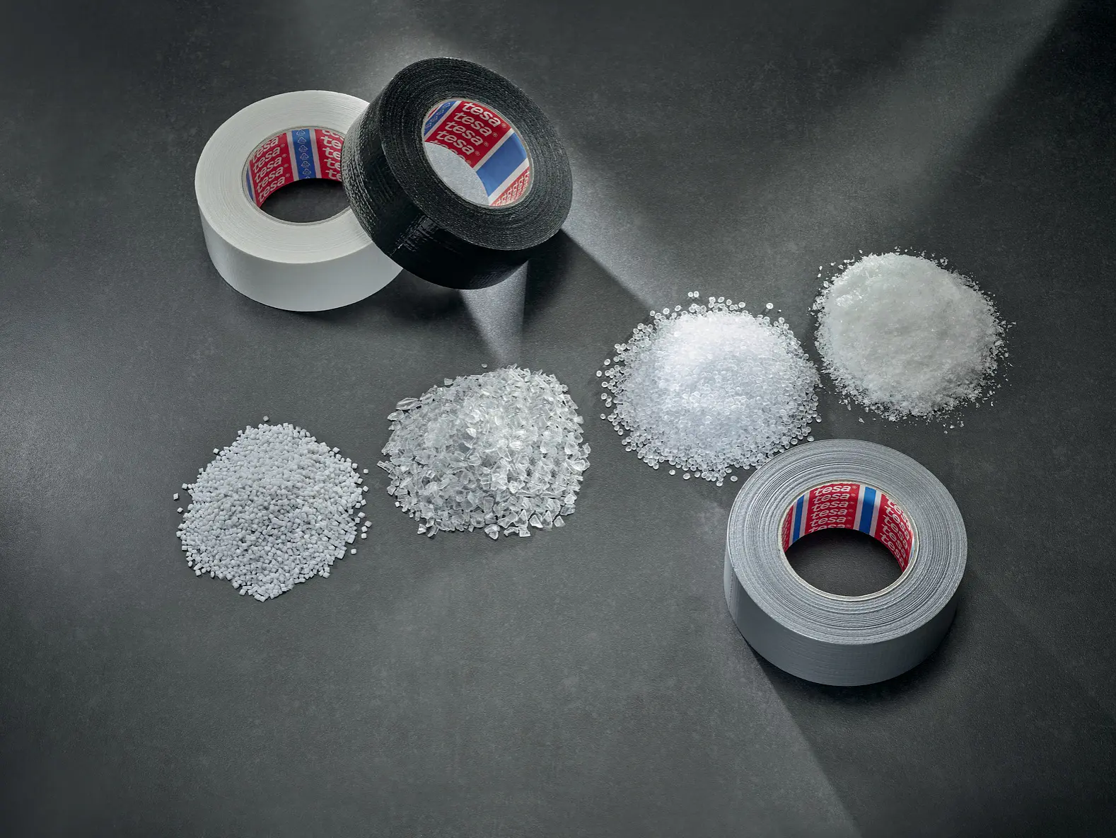 Three rolls of tesa tape—one black and two white—are displayed on a gray surface, each aligned with a small pile of plastic pellets or granules, suggesting the materials used to make the tesa tapes. (This text has been generated by AI)