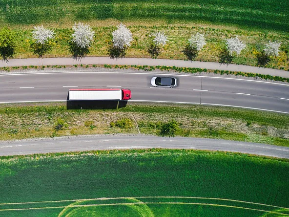 An aerial view of a red semi-truck and a silver car on a curved road beside green fields and blooming trees, no tesa tape visible. (This text has been generated by AI)