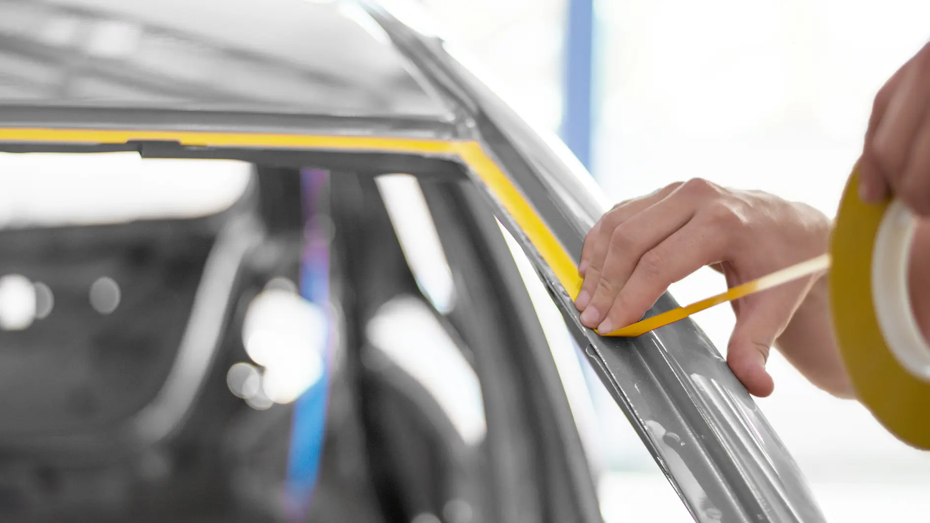 hand applying yellow masking tape along car window frame in a bright indoor auto workshop (This text has been generated by AI)