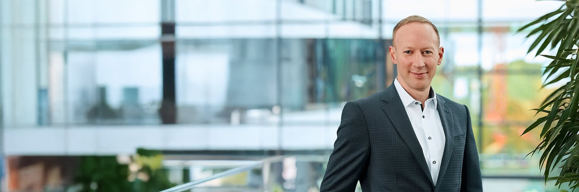 A man in a dark suit and white shirt stands indoors near a glass railing, with tesa tape visible and greenery in the background. (This text has been generated by AI)