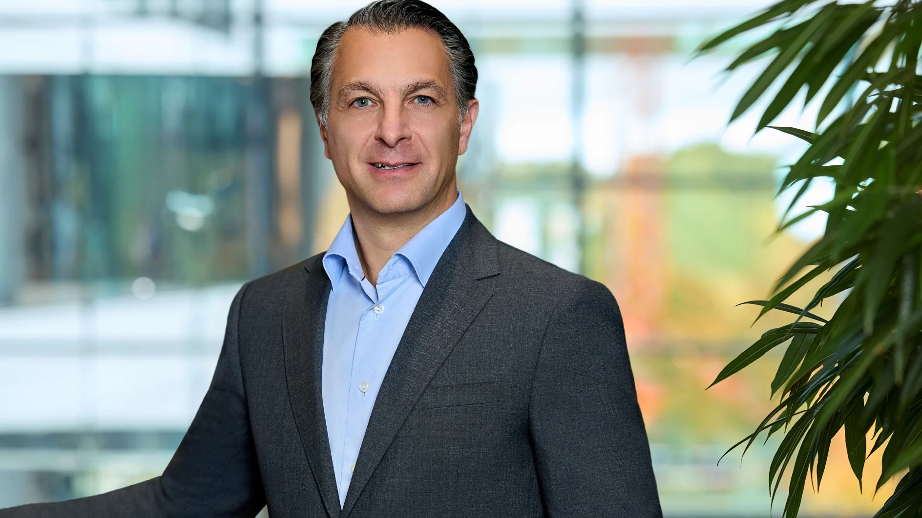 A man in a gray suit and blue shirt stands by large windows and a green plant in a modern office, smiling at the camera. (This text has been generated by AI)