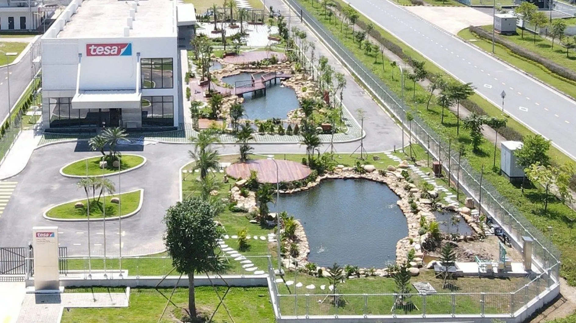 Aerial view of a modern industrial building labeled tesa tape, with landscaped grounds, ponds, palm trees, decks, and a nearby road.