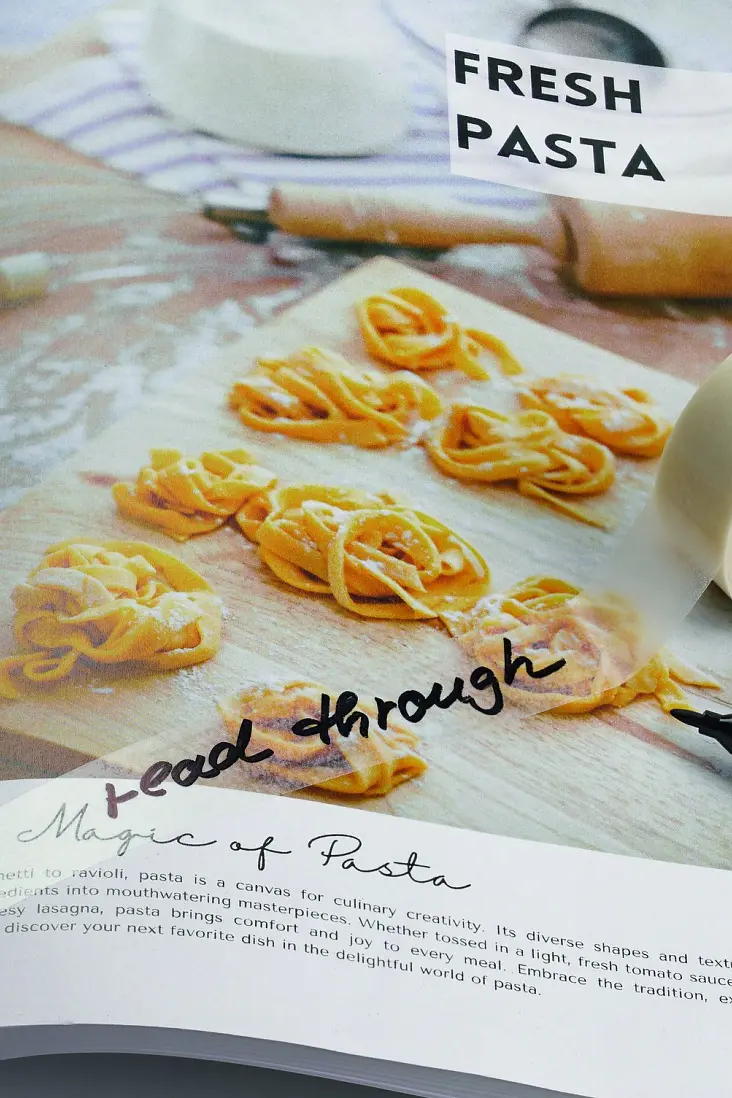 An open cookbook on a wooden surface shows pizza and pasta. A black pen and a roll of tesa tape rest on the fresh pasta page. (This text has been generated by AI)