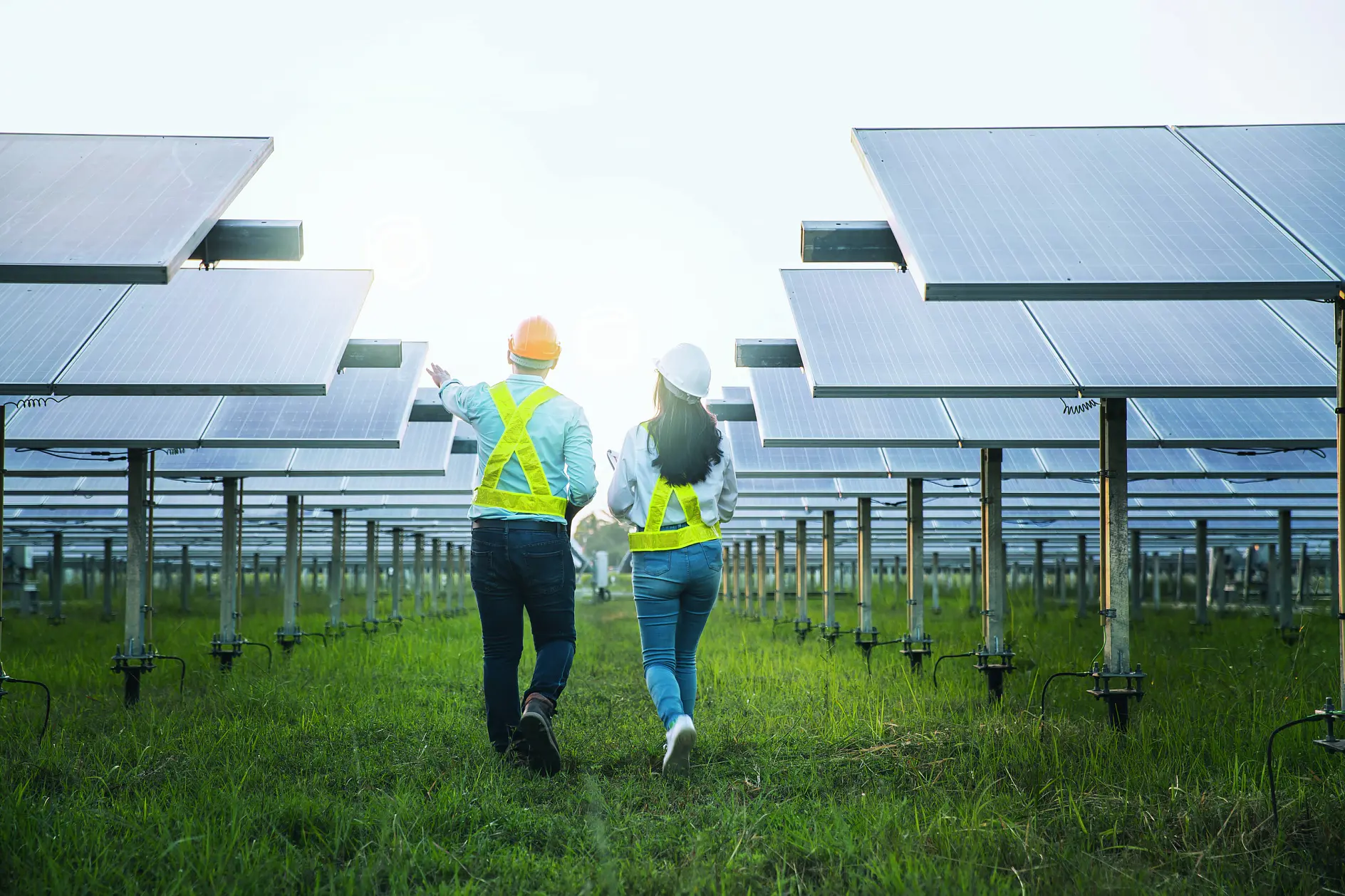 Two people wearing safety vests and helmets walk between rows of solar panels on a green field. They appear to be inspecting or discussing the solar installation. The sun is bright in the background, illuminating the panels. (This text has been generated by AI)