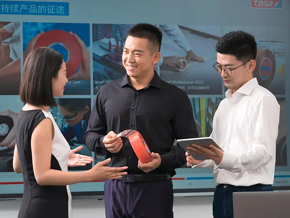 Three people stand indoors before a screen. One man holds a red roll of tesa tape; another writes on a tablet; a woman gestures. (This text has been generated by AI)