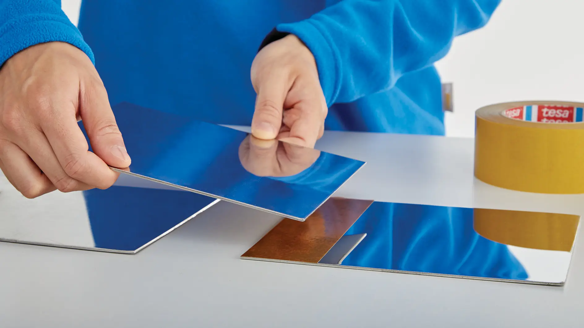 hands in blue sleeves applying clear tesa tape to a reflective surface on a white table indoors