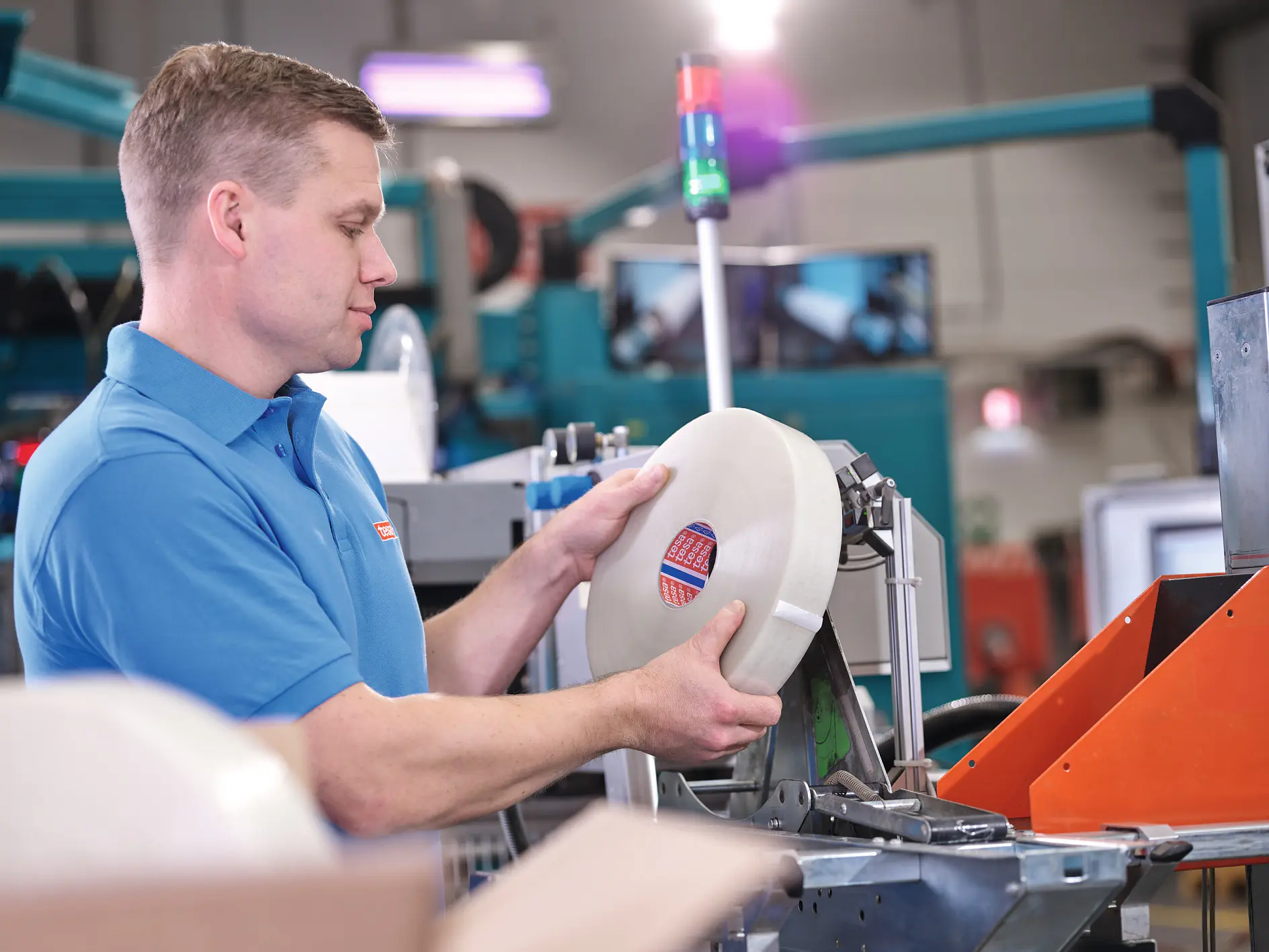 man in blue tesa shirt holding a roll of tape in an industrial setting with machinery and equipment around him