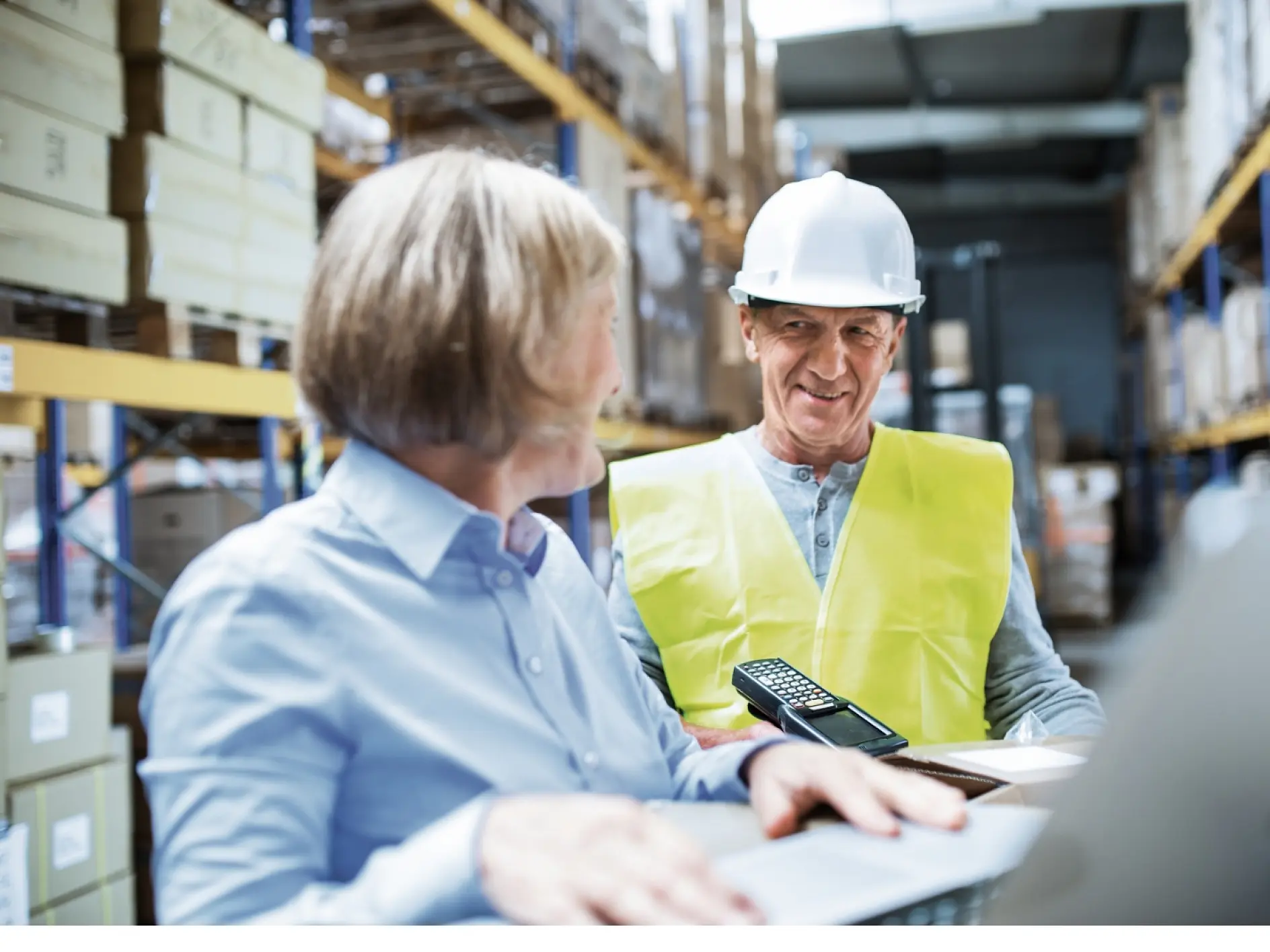 Two people in a warehouse, one wearing a safety vest and hard hat, holding a handheld scanner. They are standing near shelves with boxes sealed using tesa tape and appear to be discussing something while working.