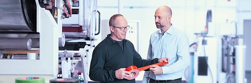 Two men stand in a factory setting holding a roll of red tesa tape. One wears a black sweater and glasses; the other is in a light blue shirt and gray pants. Multiple industrial machines and rolls of colored tesa tape are visible in the background. (This text has been generated by AI)