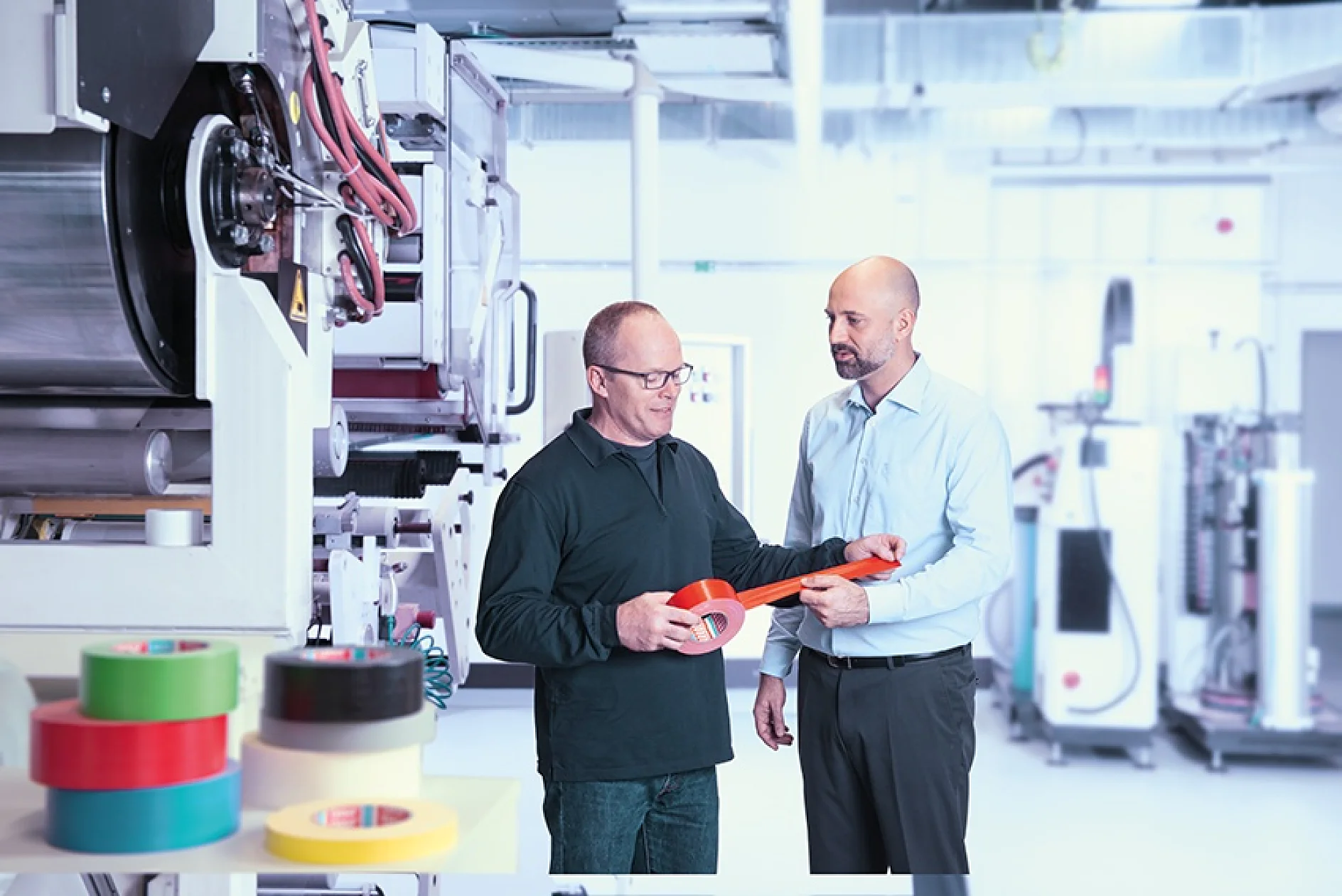 Two men stand in a factory setting holding a roll of red tesa tape. One wears a black sweater and glasses; the other is in a light blue shirt and gray pants. Multiple industrial machines and rolls of colored tesa tape are visible in the background. (This text has been generated by AI)