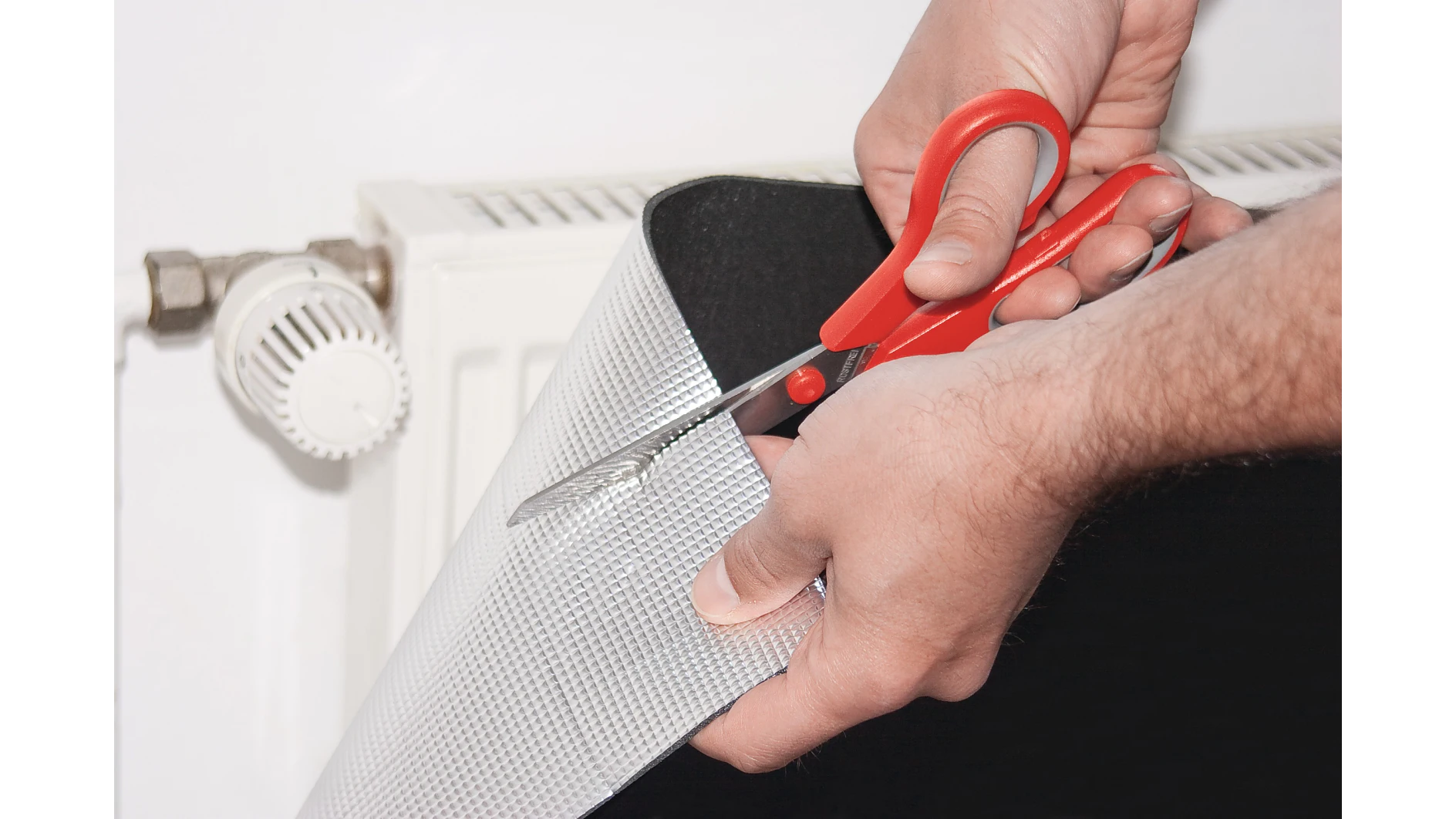 tesa_55157_ap_002_fullsize Hands cutting a silver reflective radiator insulation sheet with red scissors, with a white radiator and thermostatic valve in the background.
