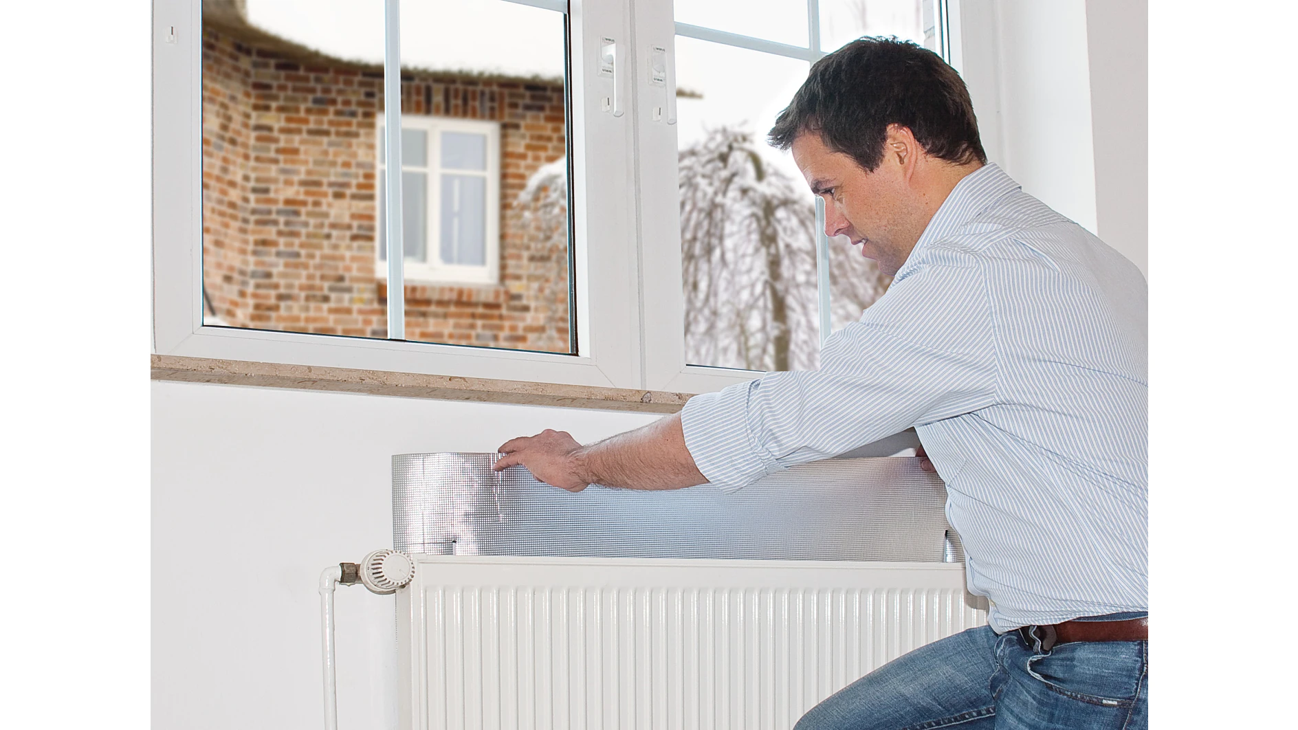 tesa_55157_ap_004_fullsize Person installing a reflective insulation sheet on a white radiator beneath a window, with a brick building visible outside.