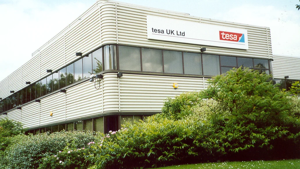 A modern two-story building with horizontal white paneling, surrounded by greenery. A sign on the upper part of the building reads tesa UK Ltd alongside the tesa logo, which is red with white letters on a blue and white background. (This text has been generated by AI)