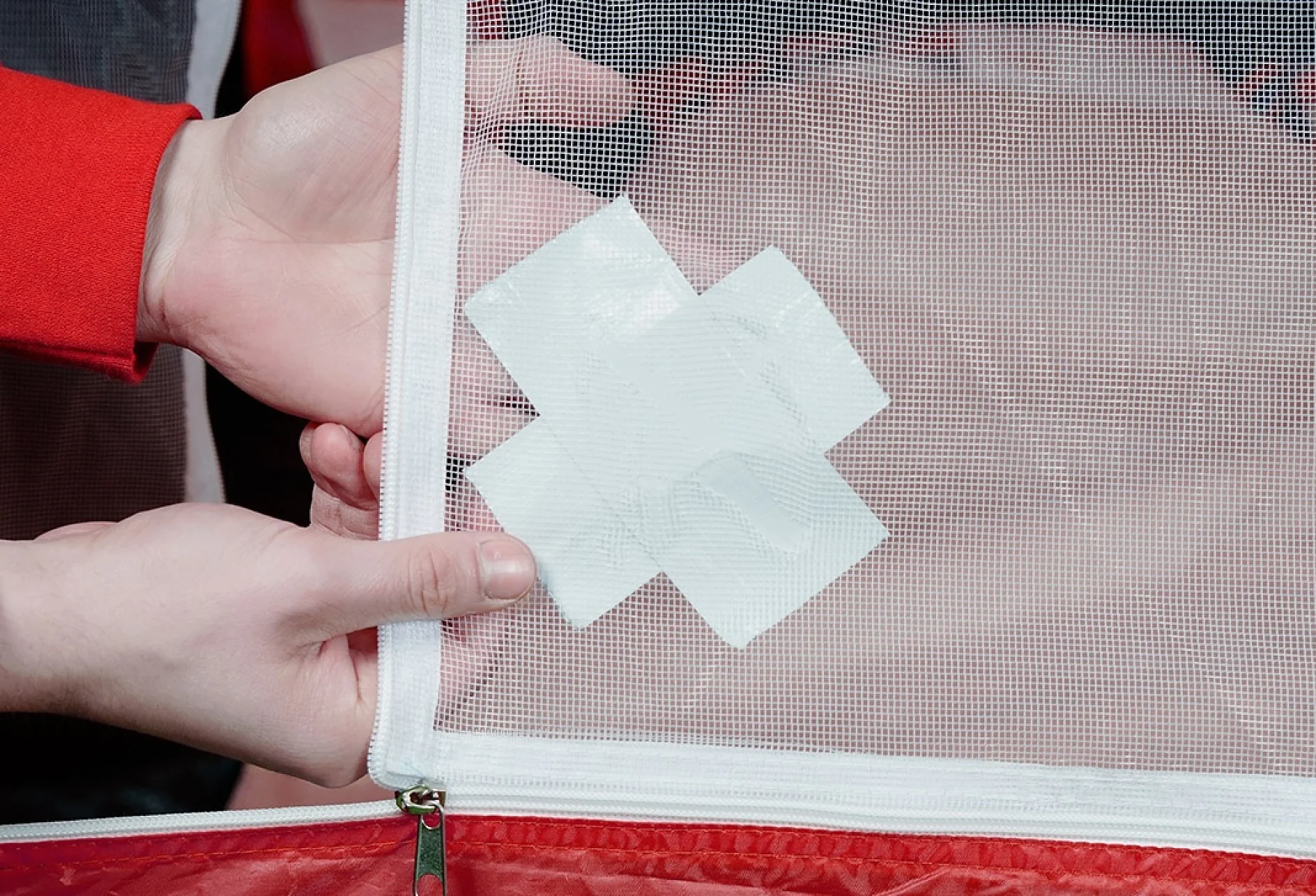 Two hands hold a mesh fabric with a white, cross-shaped patch of tesa tape on it, set against a red base. (This text has been generated by AI)
