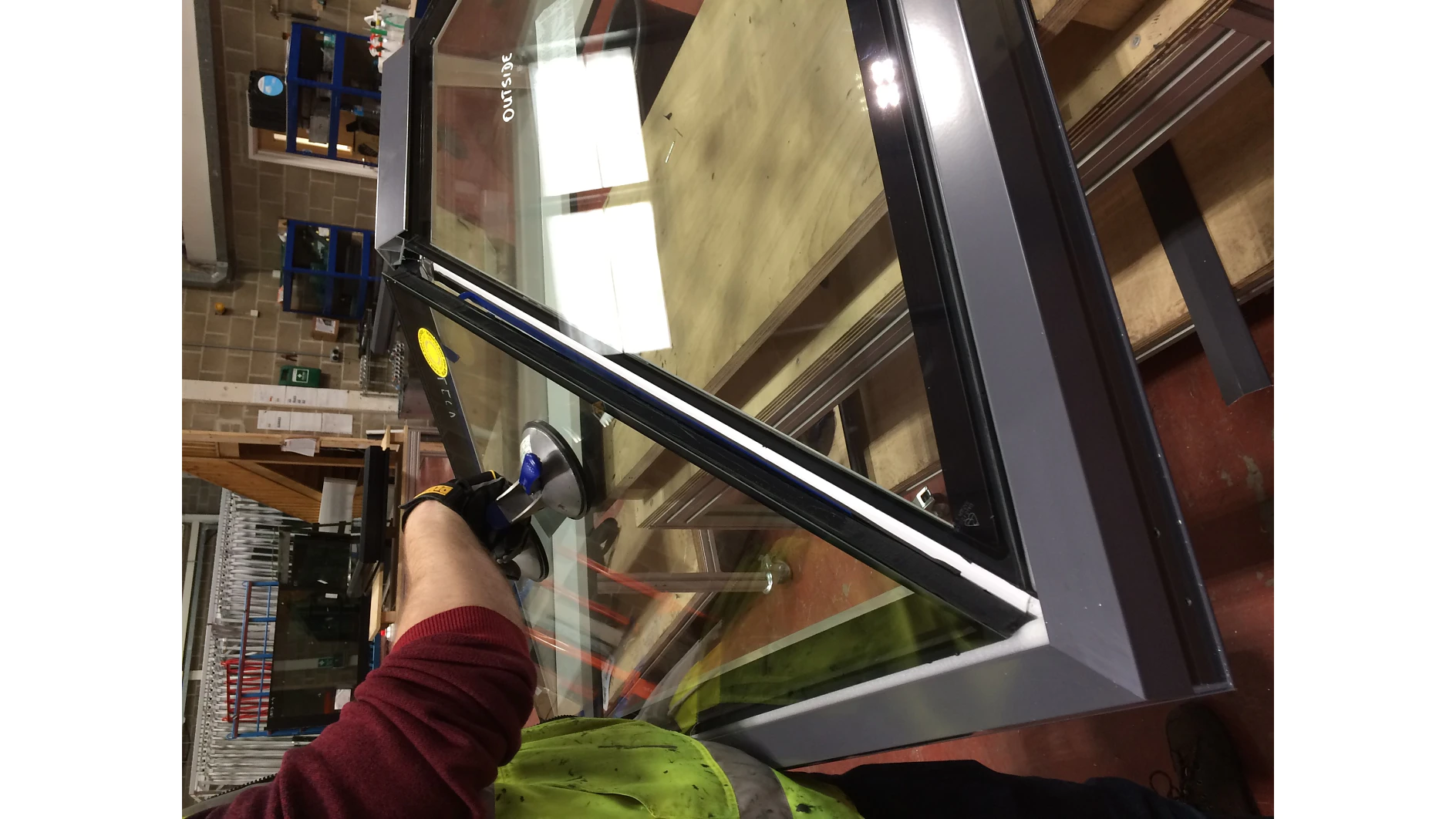 A person in a green safety vest is holding a large glass window with black and yellow suction cup handles. The window is tilted, and various tools, including tesa tape, are visible in the background. (This text has been generated by AI)