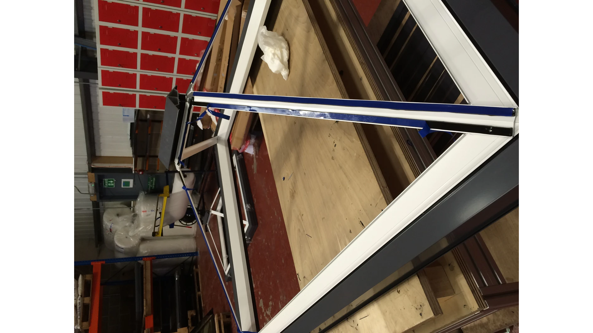 A large rectangular window frame partially wrapped in protective tesa tape lies on a wooden table in a workshop. Shelves with red boxes and tools, along with a fire extinguisher, are visible in the background. (This text has been generated by AI)