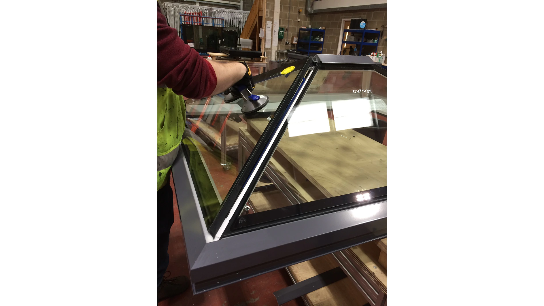 A person is using a suction tool to handle a large piece of triangular glass. They are wearing a glove on their right hand, and the panel is secured with tesa tape. The glass panel is surrounded by a gray metal frame and is situated in an indoor workshop environment. (This text has been generated by AI)