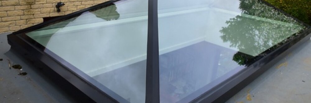 A large glass skylight with a black metal frame is installed on a flat roof. It has a pyramid shape and provides a view of the room below, with books visible inside. The background shows a brick wall and greenery. (This text has been generated by AI)