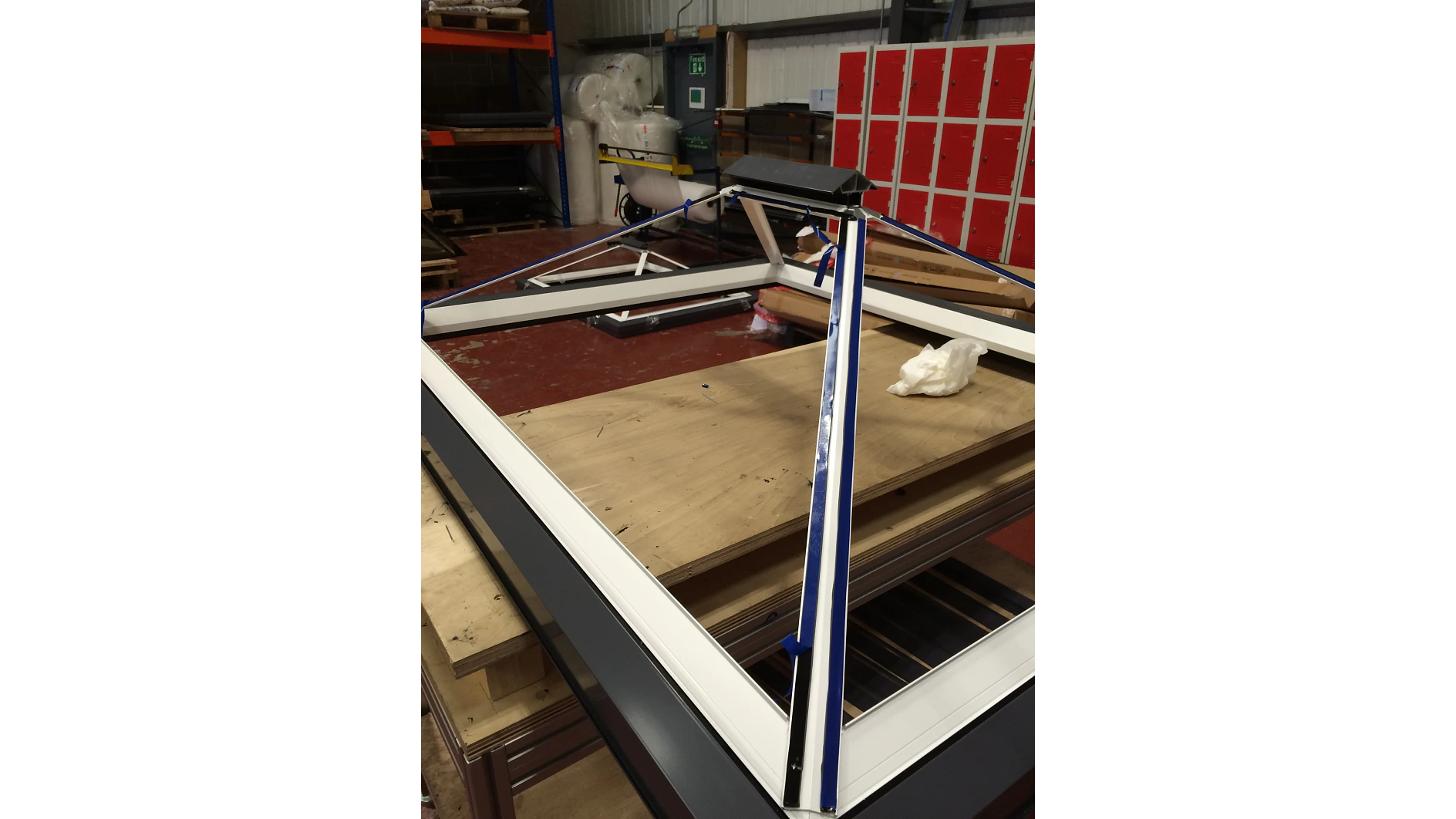 A large, angular metal frame rests on a workbench in a workshop. The frame has blue tesa tape strips along its edges. In the background, there are red lockers, stacked materials, and various industrial equipment. (This text has been generated by AI)
