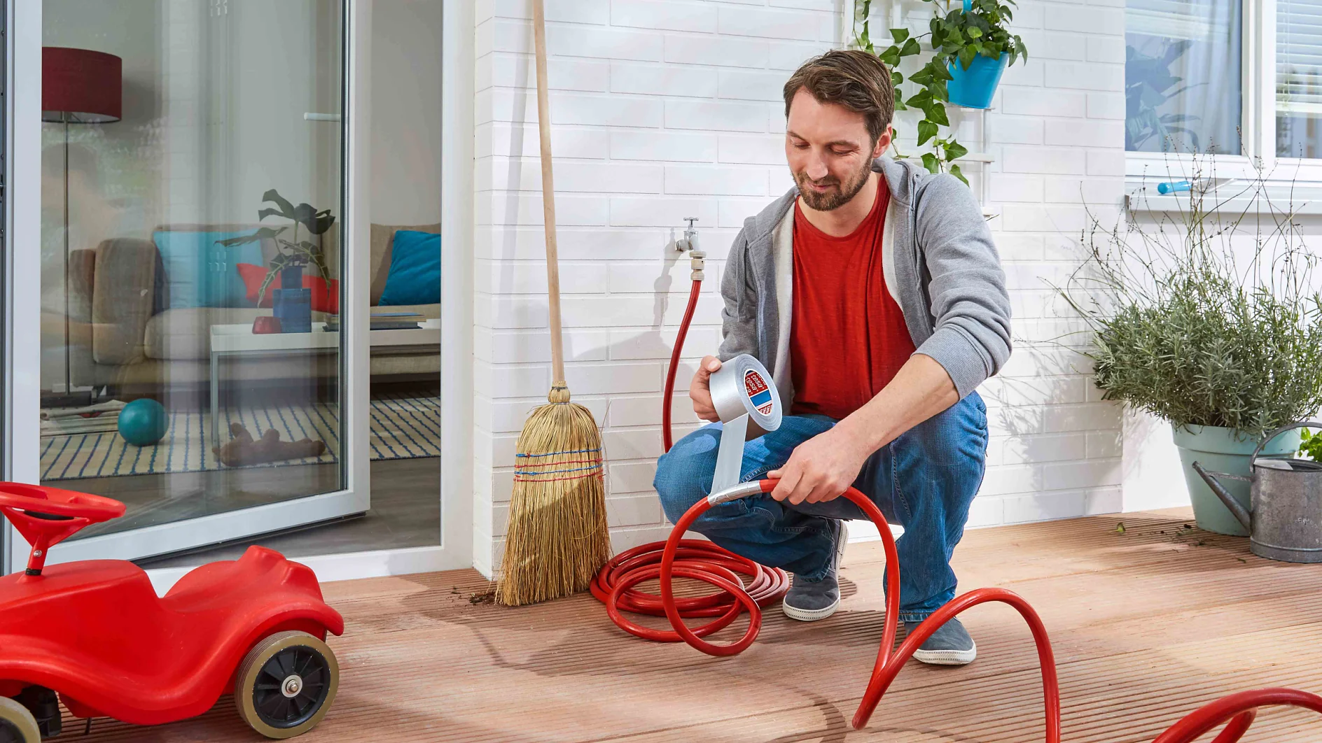A man kneels on a wooden deck connecting a red garden hose to a faucet. Near him, there is tesa tape securing the connection. A red toy car is nearby. The interior of a house is visible through a glass door in the background. A broom leans against the wall, and there are plants around. (This text has been generated by AI)