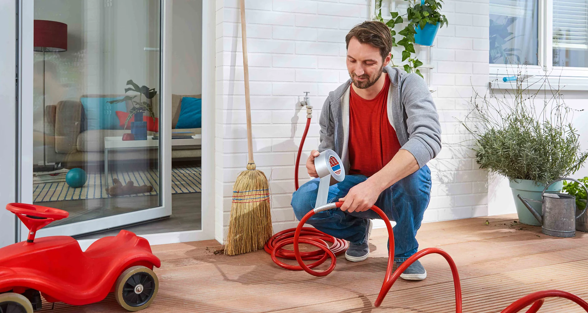 A man kneels on a wooden deck connecting a red garden hose to a faucet. Near him, there is tesa tape securing the connection. A red toy car is nearby. The interior of a house is visible through a glass door in the background. A broom leans against the wall, and there are plants around. (This text has been generated by AI)