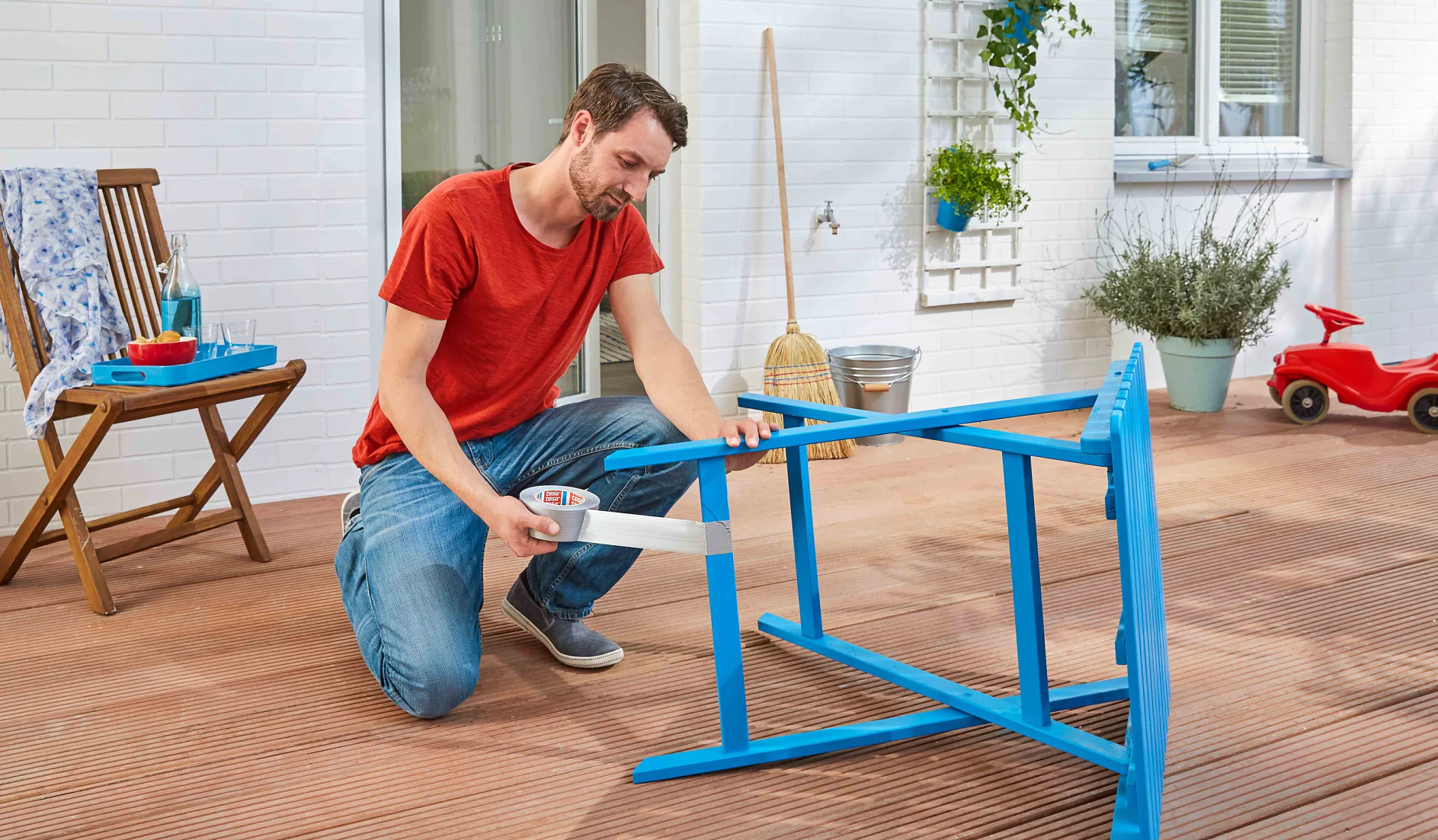 A man in a red shirt and jeans kneels on a wooden deck, using a handheld vacuum to clean a blue wooden chair turned on its side. A broom, potted plants, and a red toy car are in the background. (This text has been generated by AI)