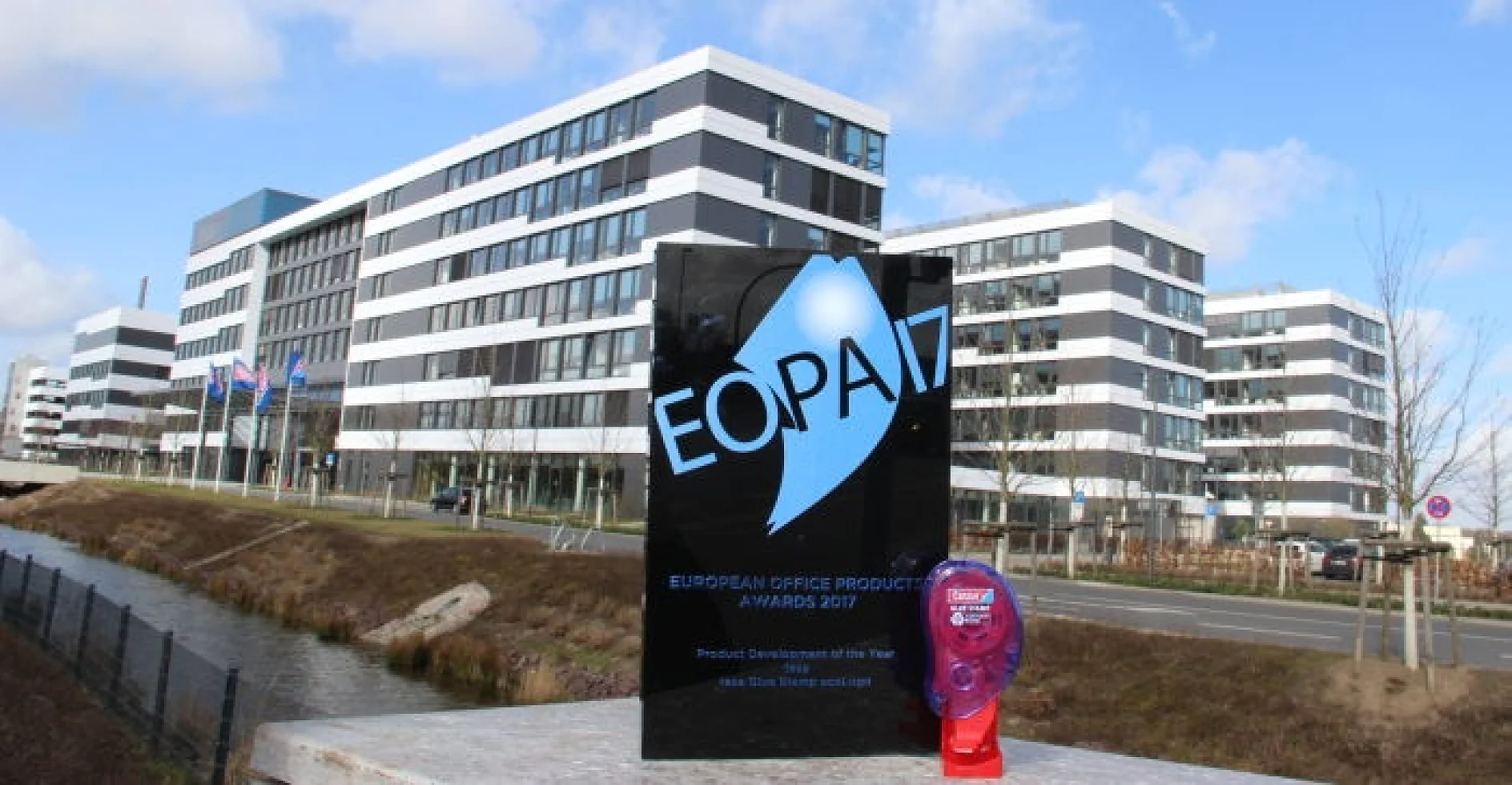 A black award plaque with white and blue text reading EOPA 17 stands on a ledge next to a small purple fan with a circular red badge. In the background, there is a modern multi-story office building and a road. Every element holds its position securely, thanks to the reliable grip of tesa tape. (This text has been generated by AI)