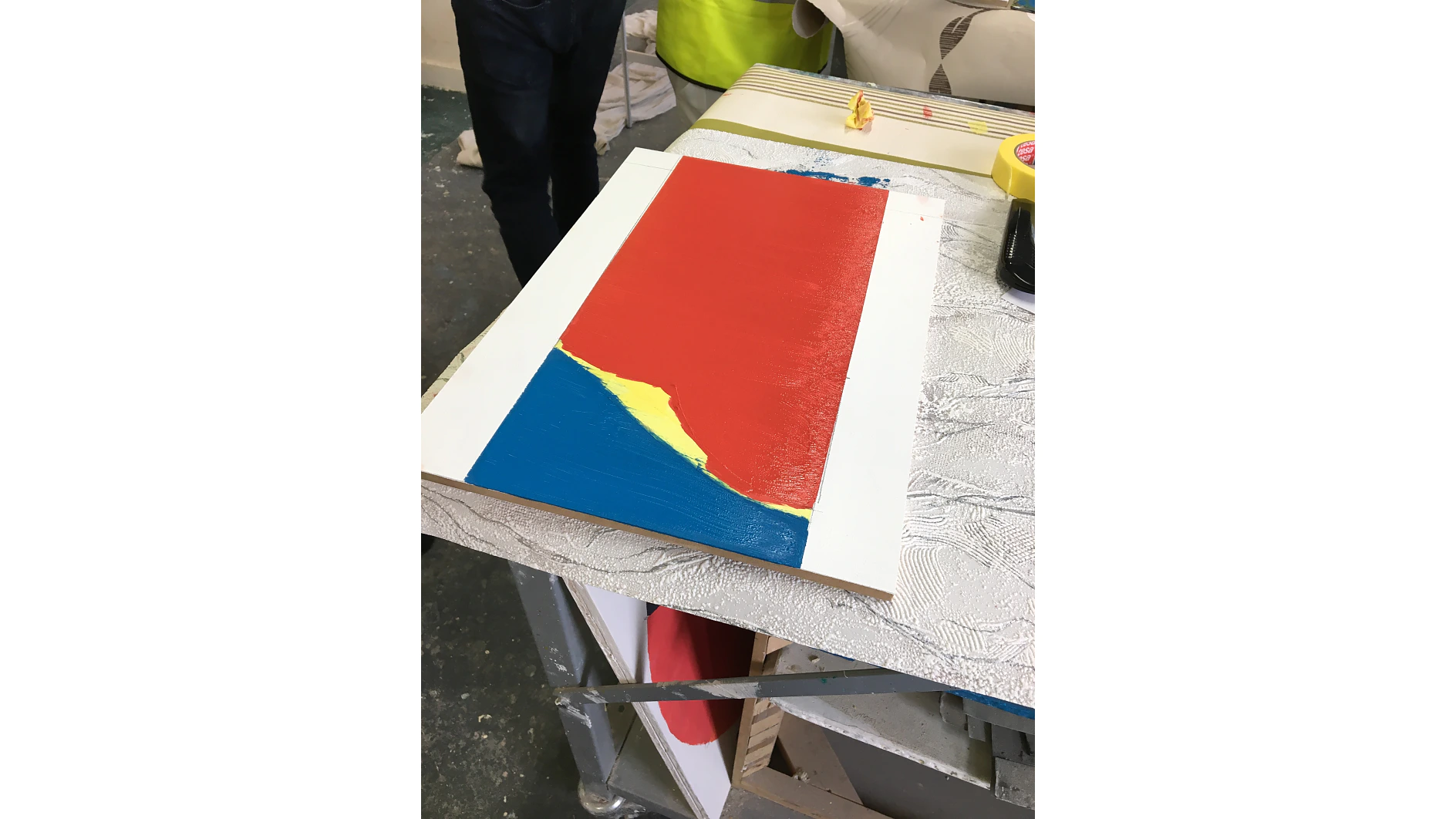 A rectangular canvas on a table partially painted with bold red, blue, and yellow colors. A person wearing black pants and shoes stands nearby. Paint supplies and rolls of tesa tape are visible in the background. (This text has been generated by AI)