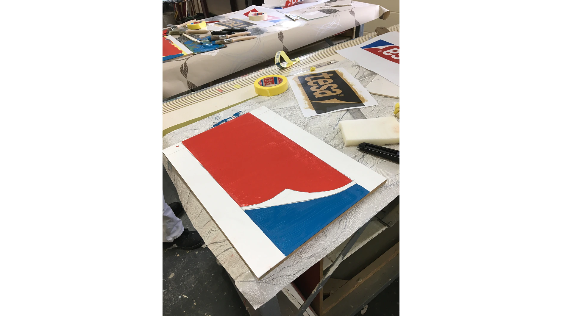 A worktable with art supplies, including tesa tape, paint containers, and paintbrushes. A partially completed painting on paper features red, blue, and white sections. A stencil with the word Pepsi is visible in the background. (This text has been generated by AI)