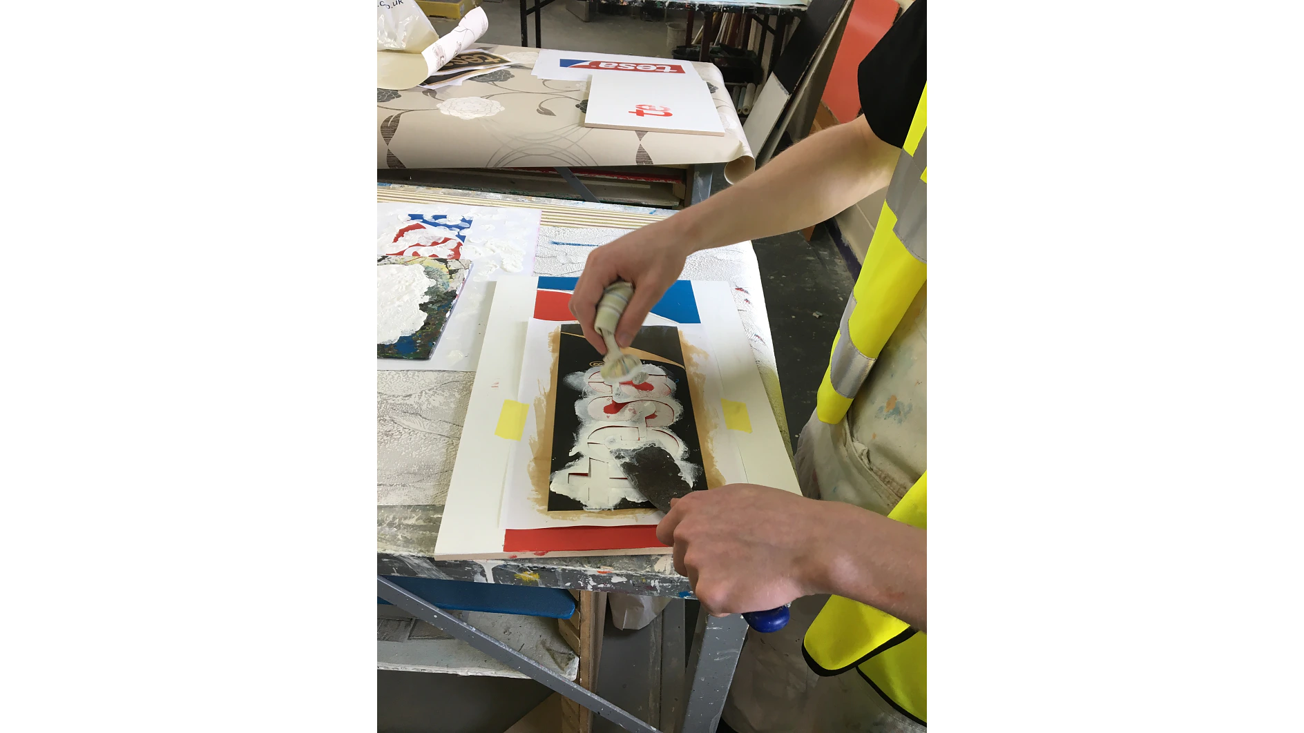 A person wearing a yellow vest spray-paints a stencil with layers of red and white paint on a table. Other art supplies, papers, and rolls of tesa tape are visible in the background. (This text has been generated by AI)