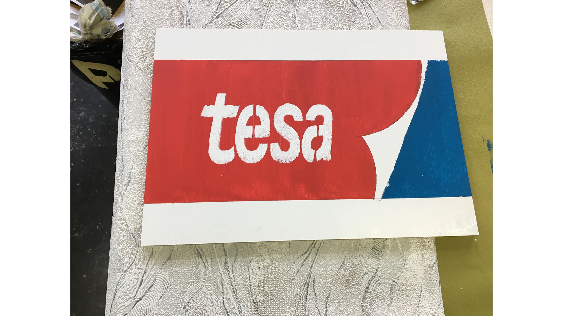 A rectangular signage with the word tesa in white lowercase letters on a red background. On the right, theres a blue section partially covered by a red curved shape. The surface beneath is textured and light-colored. (This text has been generated by AI)