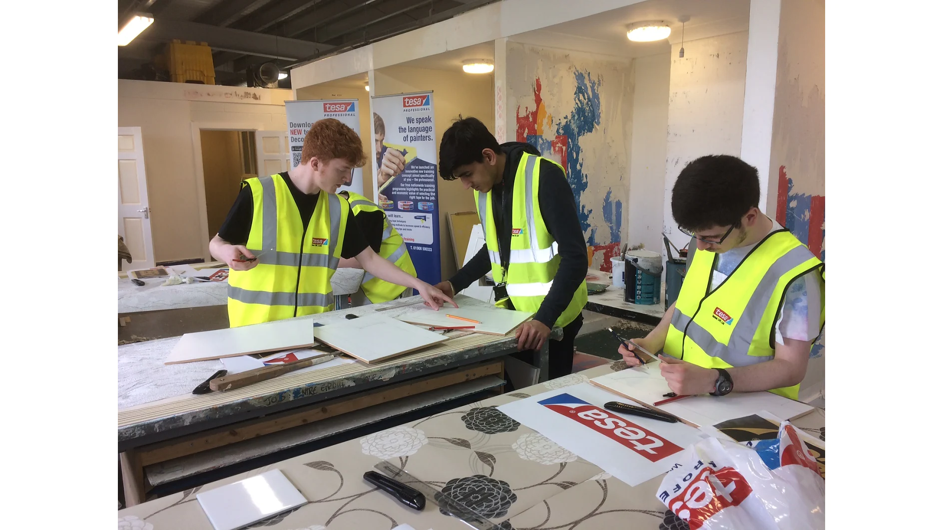 Three individuals wearing high-visibility vests work on a project in a workshop. They are focused on measuring and cutting materials on tables, using tesa tape for precision. The environment is industrial, with tools and equipment visible in the background. (This text has been generated by AI)