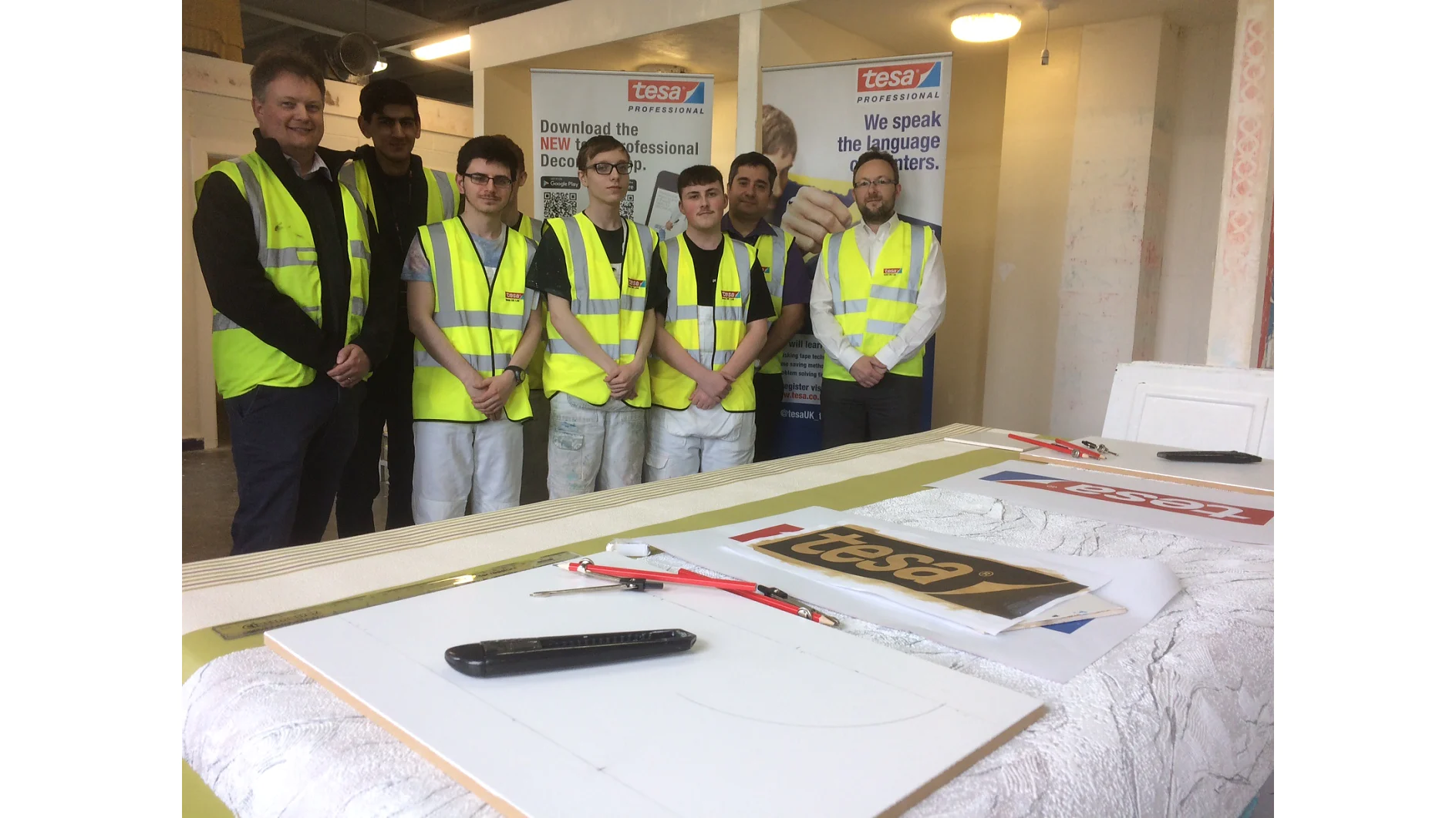 Seven people wearing yellow safety vests stand in a row in front of banners displaying tesa professional. A table in the foreground holds various tools, papers, and a box with the tesa logo. The setting appears to be a workshop or training space. (This text has been generated by AI)