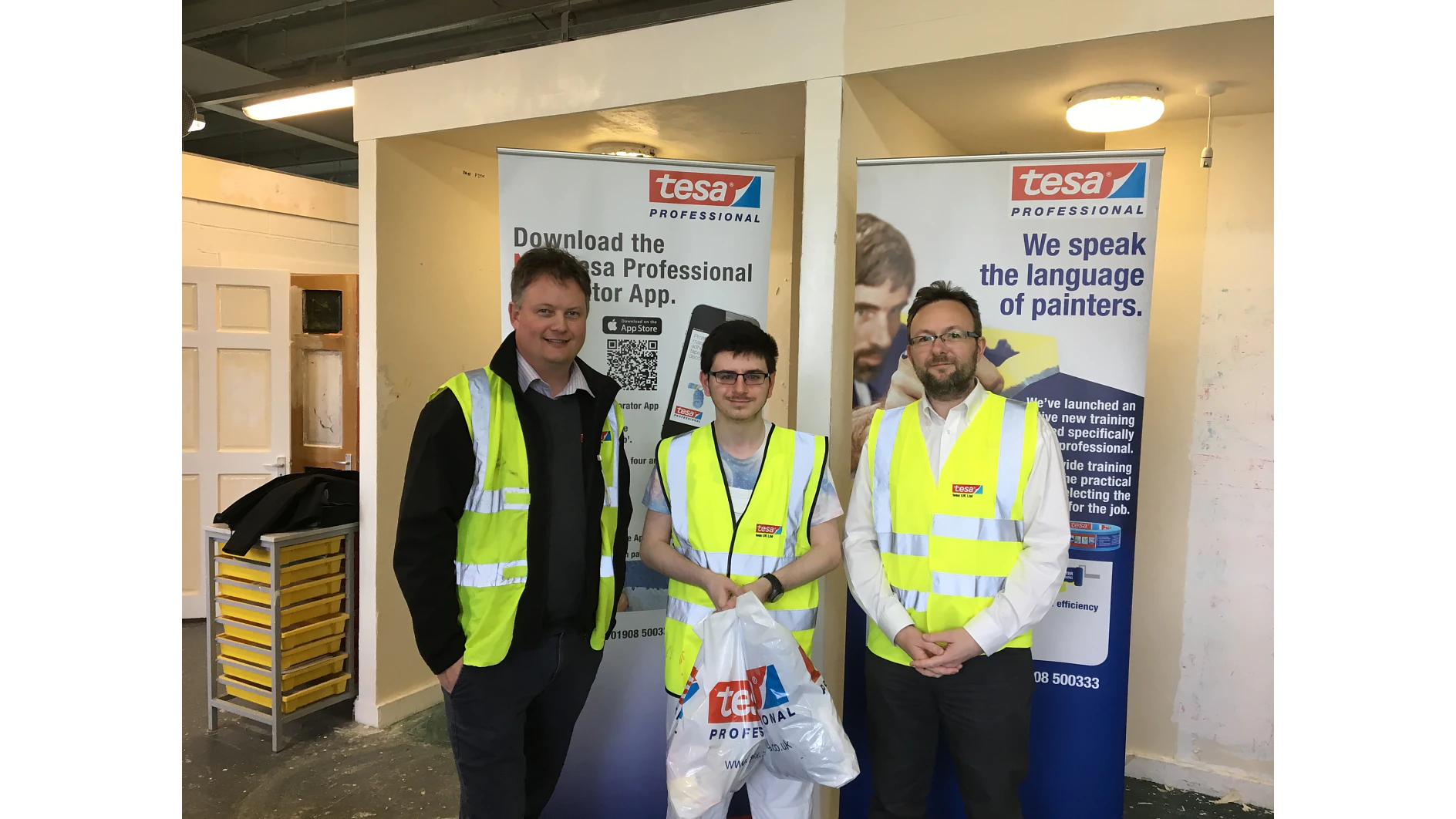 Three individuals in high-visibility vests stand indoors in front of two banners displaying tesa Professional logos. One person is holding a plastic bag with the same logo. The background shows a partially visible doorway and interior wall. (This text has been generated by AI)